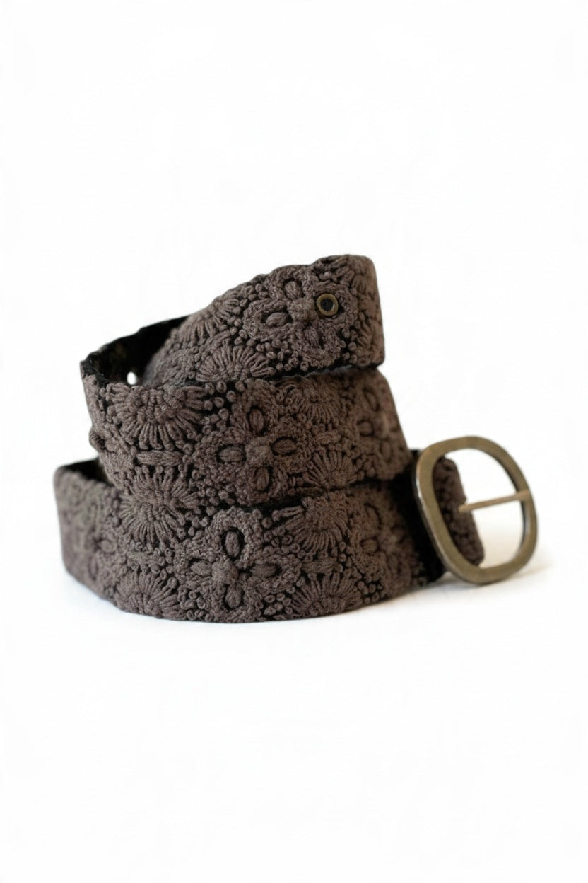 The JENNY KRAUSS - MIDNIGHT OASIS BELT features intricate raised floral embroidery, a round metal buckle, and a dark brown design, handcrafted in Peru and shown coiled on a light background.