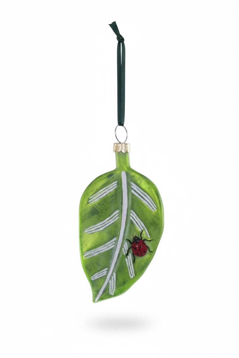 The CODY FOSTER GARDEN FRIENDS ORNAMENT is a charming glass leaf with white veins, a red ladybug detail, and a dark green hanging ribbon—perfect for any nature lover’s collection.