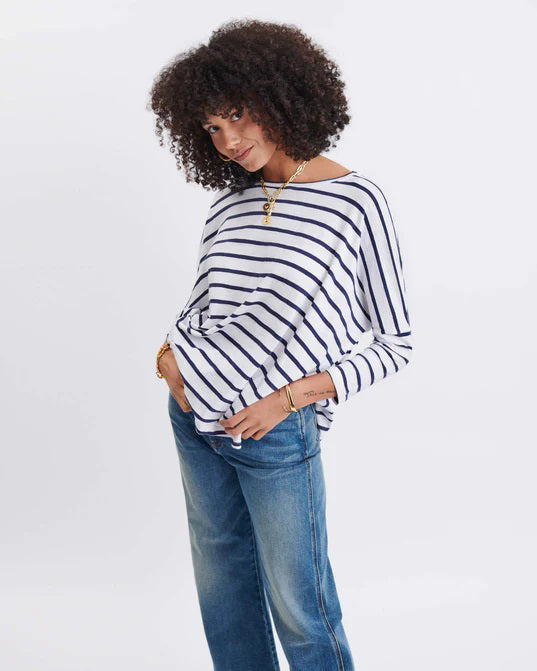 A woman with long, straight hair stands against a white wall, wearing the MERSEA Catalina Slub Tee in blue paired with white pants. The midweight textured cotton tee is from MERSEA. She looks at the camera with a neutral expression.