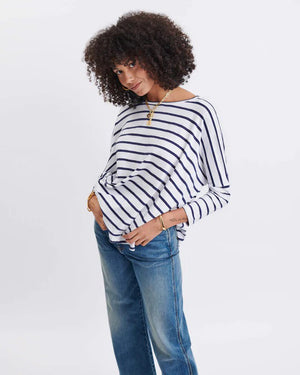 A woman with long, straight hair stands against a white wall, wearing the MERSEA Catalina Slub Tee in blue paired with white pants. The midweight textured cotton tee is from MERSEA. She looks at the camera with a neutral expression.
