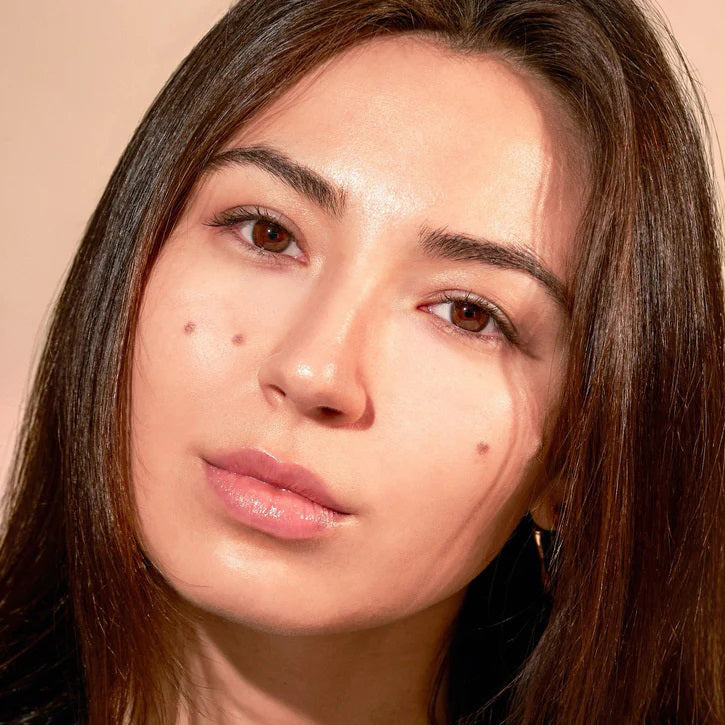 A close-up of a person with natural curly hair, adorned in gold hoop earrings and a white top. They wear a neutral facial expression while looking directly at the camera. The softly lit, plain background enhances their glowing skin as if touched by JASON WU - TINTED MOISTURIZER MEETS CC CREAM from JASON WU BEAUTY.