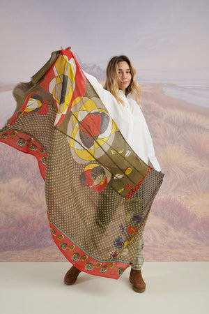 A woman with long blonde hair smiles at the camera in a scenic setting, wearing a white shirt, plaid pants, and the STORIATIPIC - ASTER WOOL SCARF by STORIATIPIC. Mountains, grass, and water are visible in the background.