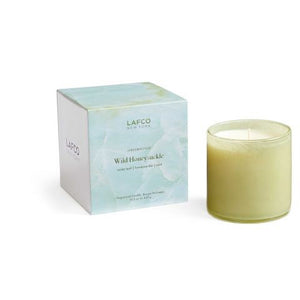 A light green soy wax candle in a hand-blown glass container is placed next to its packaging box. The box, with a watercolor design featuring shades of green and white, displays the text "LAFCO New York, Greenhouse, Wild Honeysuckle.