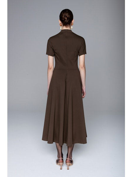 A woman faces away, wearing the LAVI COUTURE - MIDI DEIOPEA DRESS by LAVI in brown. The minimalist design features short sleeves and a fitted waist, styled with a neat bun and heeled sandals against a plain gray background.