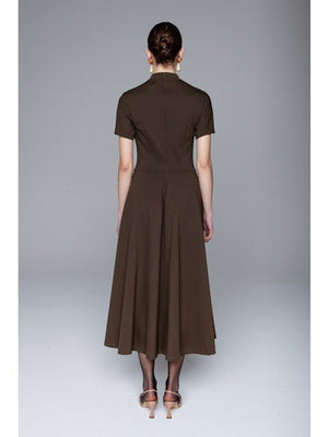 A woman faces away, wearing the LAVI COUTURE - MIDI DEIOPEA DRESS by LAVI in brown. The minimalist design features short sleeves and a fitted waist, styled with a neat bun and heeled sandals against a plain gray background.