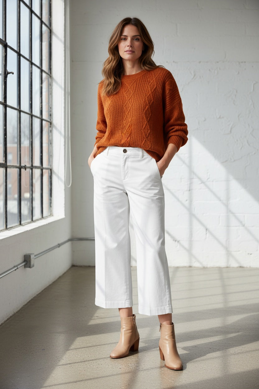 A person stands indoors by large windows, wearing a textured orange sweater, BRAX - MAINE WIDE LEG CULOTTE IN WHITE by BRAX, and tan ankle boots. Hands are in pockets. Natural light casts soft shadows on the floor and white brick wall.