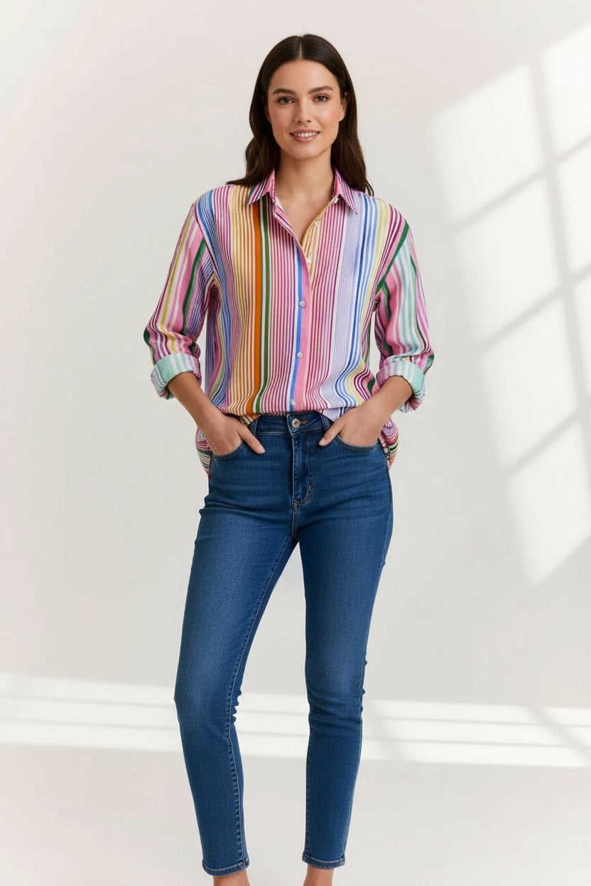 A woman in the HINSON WU Halsey Long Sleeve Linen Shirt in Multi Stripe, featuring vibrant vertical stripes, rolled-up sleeves, and lightweight linen fabric, pairs it with blue jeans. She has straight brown hair, hoop earrings, and a bracelet.