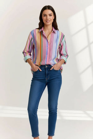 A woman in the HINSON WU Halsey Long Sleeve Linen Shirt in Multi Stripe, featuring vibrant vertical stripes, rolled-up sleeves, and lightweight linen fabric, pairs it with blue jeans. She has straight brown hair, hoop earrings, and a bracelet.