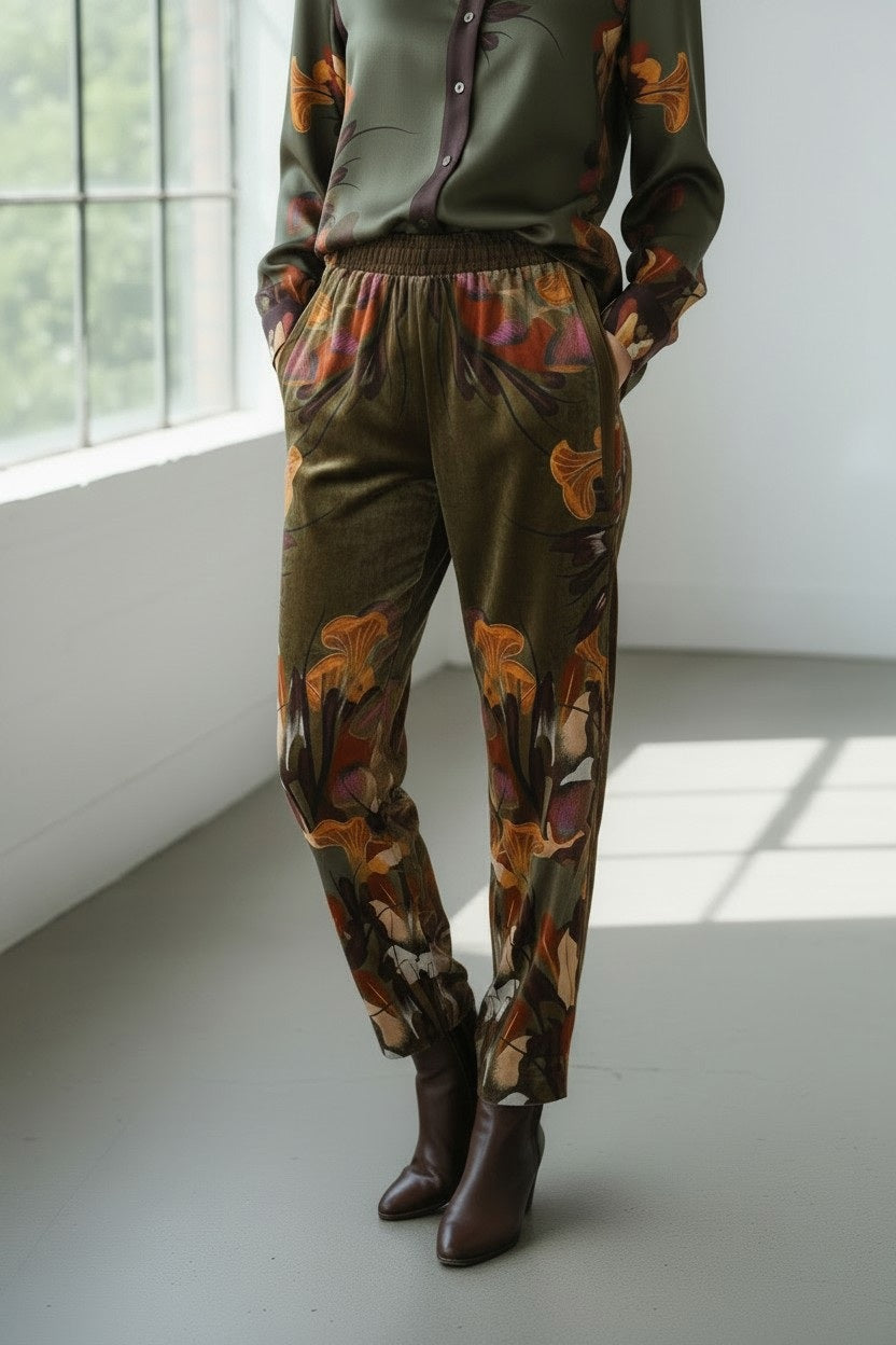 A person stands indoors in sunlight streaming through a large window to the left, wearing a green blouse and the FRANCO FERRARI - KANADA VELVET TROUSER featuring a botanical print and an elasticized waistband, hands tucked in pockets, paired with brown ankle boots.