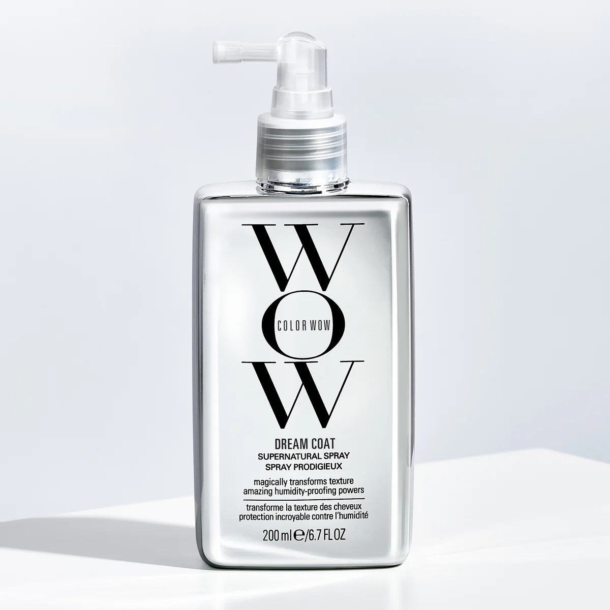 A bottle of KT SUPPLY COLORWOW - DREAM COAT SUPERNATURAL SPRAY, featuring a pump dispenser and black and white text on a shiny silver background, is placed on a light surface with soft shadowing.