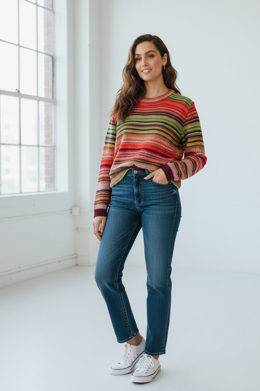 A woman stands in a bright room, smiling at the camera with one hand in her pocket, wearing the FLOOR - COTTON STRIPED SWEATER by FLOOR, blue jeans, and white sneakers.