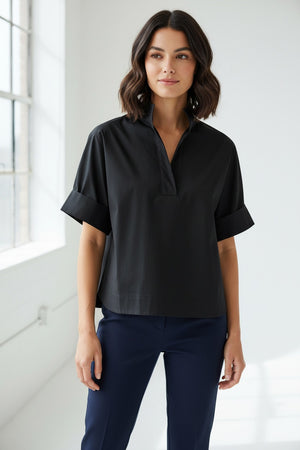 A woman with long red hair wears the HINSON WU Cara Elbow Sleeve Shirt in Stretch Polished Cotton Black—an oversized, loose-fit blouse by HINSON WU—paired with navy pants and dangling blue earrings, standing against a plain light background.