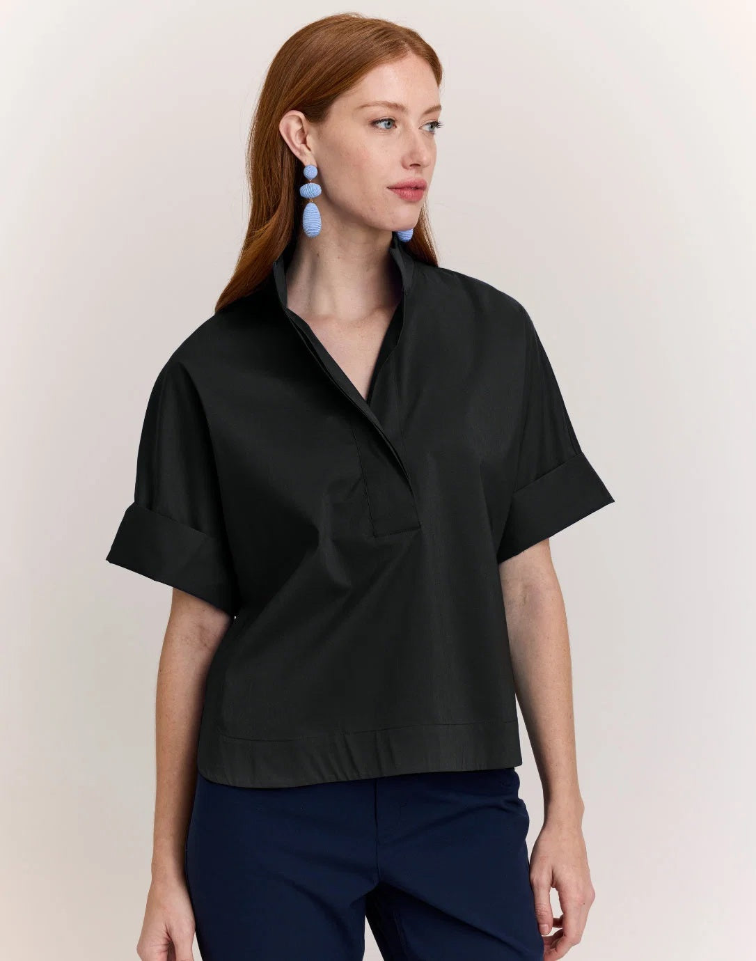 A woman with long red hair wears the HINSON WU Cara Elbow Sleeve Shirt in Stretch Polished Cotton Black—an oversized, loose-fit blouse by HINSON WU—paired with navy pants and dangling blue earrings, standing against a plain light background.