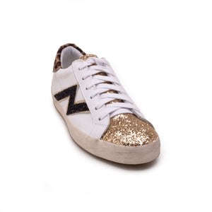 The MICHELE LOPRIORE - NINA SNEAKER is crafted from soft leather and features white laces, a silver glitter-covered toe cap, and a beige and black zigzag design on the side. It includes an extra light rubber sole with a height of 2.5 cm, a slightly worn midsole, and a black heel tab. Displayed against a plain white background, this stylish sneaker seamlessly blends elegance with contemporary flair.