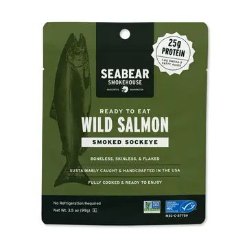 A green SEABEAR SMOKEHOUSE package of Ready-To-Eat Smoked Sockeye Salmon, featuring a salmon illustration and labels indicating 25g protein, boneless, skinless, flaked, and sustainably caught in the USA.