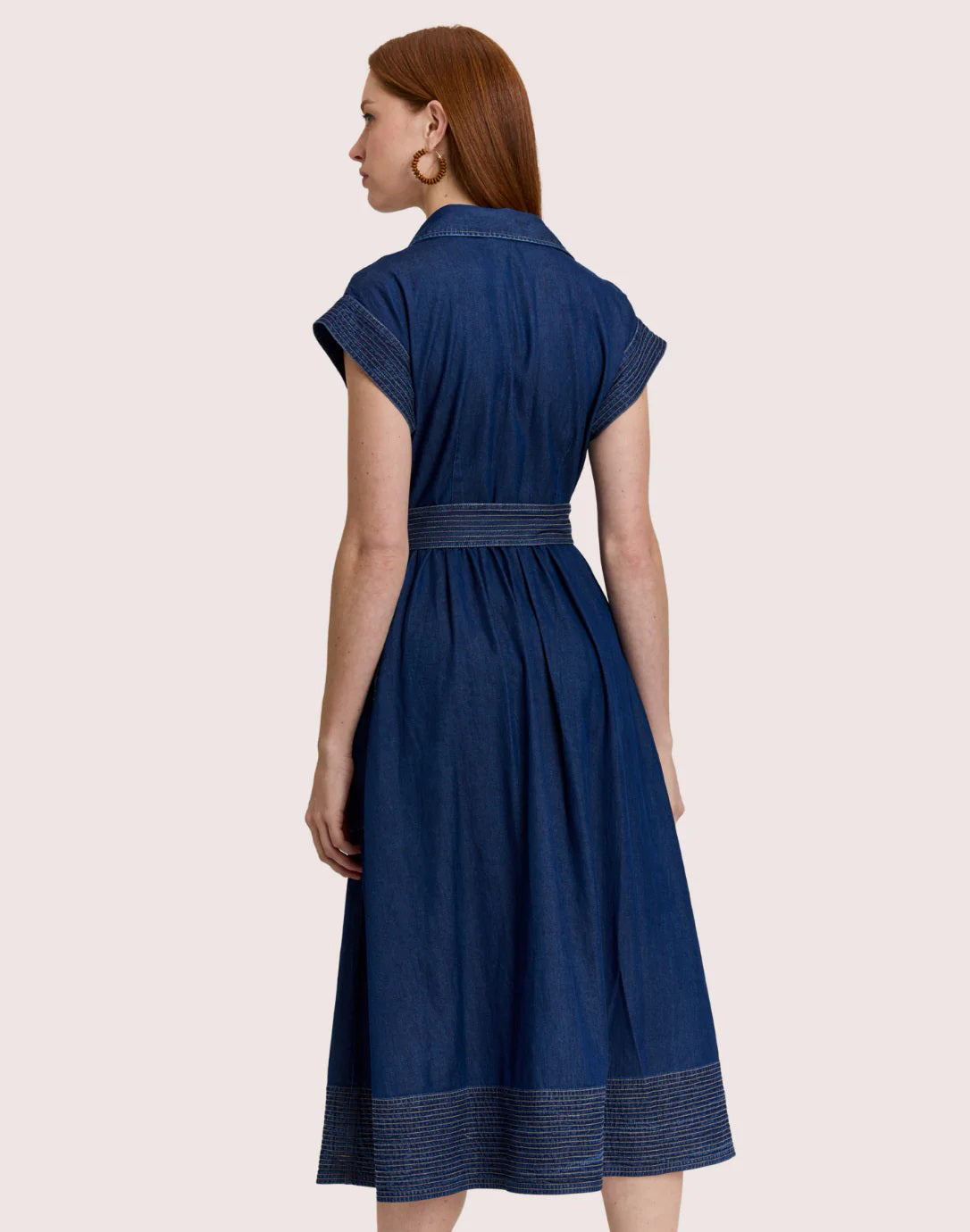 A woman stands against a light background wearing the HINSON WU Annabelle Short Sleeve Dark Wash Denim Dress, featuring a collar, button-down front, belt, pockets, and decorative stitching at the hem and sleeves.