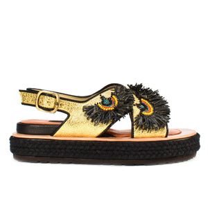 A person wearing MEHER KAKALIA's Rio Bloc Sandal in Charm Gold, featuring intricate, peacock feather-like designs and unmatched artisanal quality. With painted toenails and rolled-up jeans, they sit on a wooden floor.