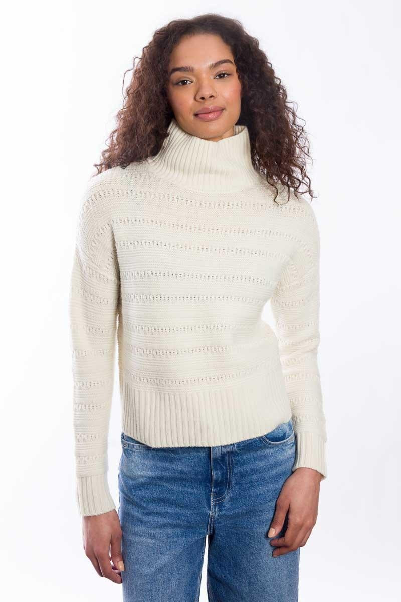 A person with curly hair wears the & ISLA Textured Mock Neck Pullover in Ivory and blue jeans, standing against a plain white background, looking confidently at the camera.