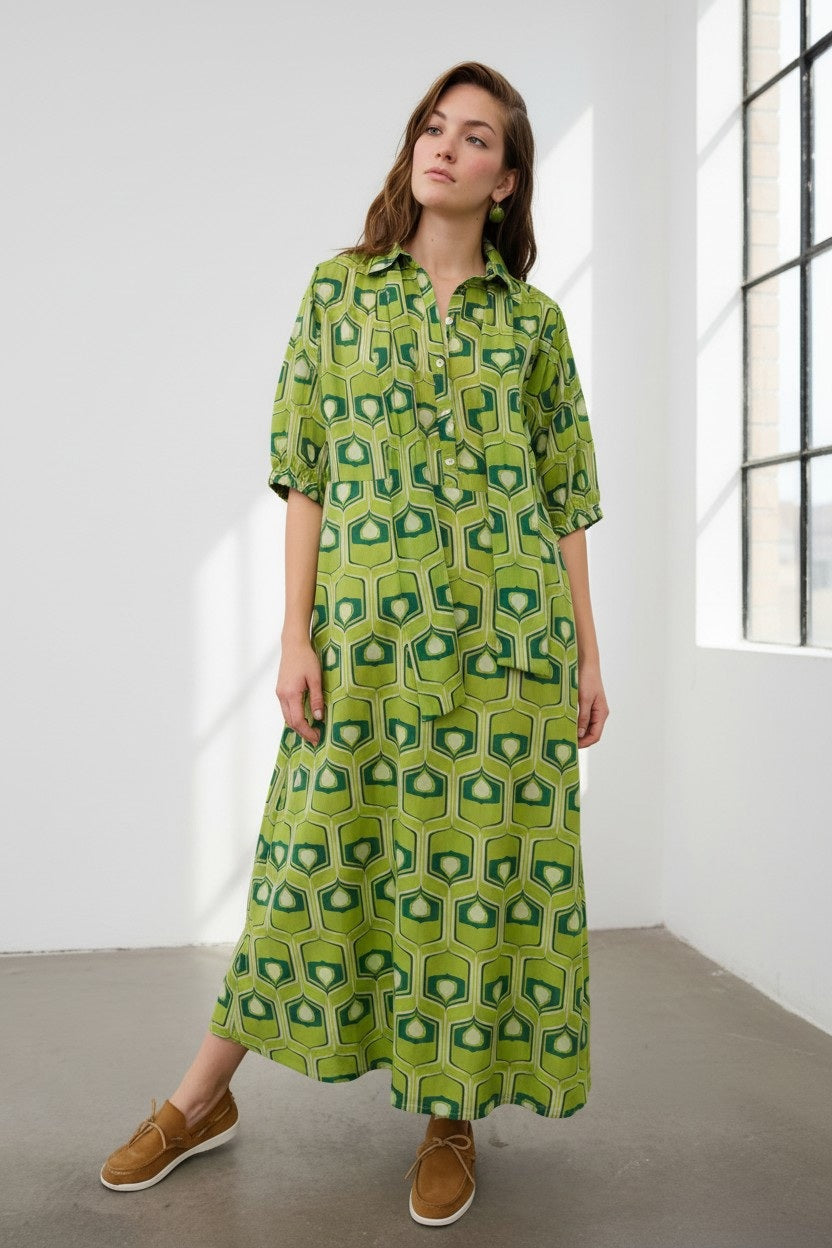 A woman stands indoors wearing the FLOOR - COTTON MAXI DRESS by FLOOR, featuring a long silhouette with a bold green geometric print skirt and paired with brown shoes. She gazes slightly upward, one hand resting by her side, as sunlight from large windows illuminates the simple floor and highlights the Italian-made elegance of this ensemble.