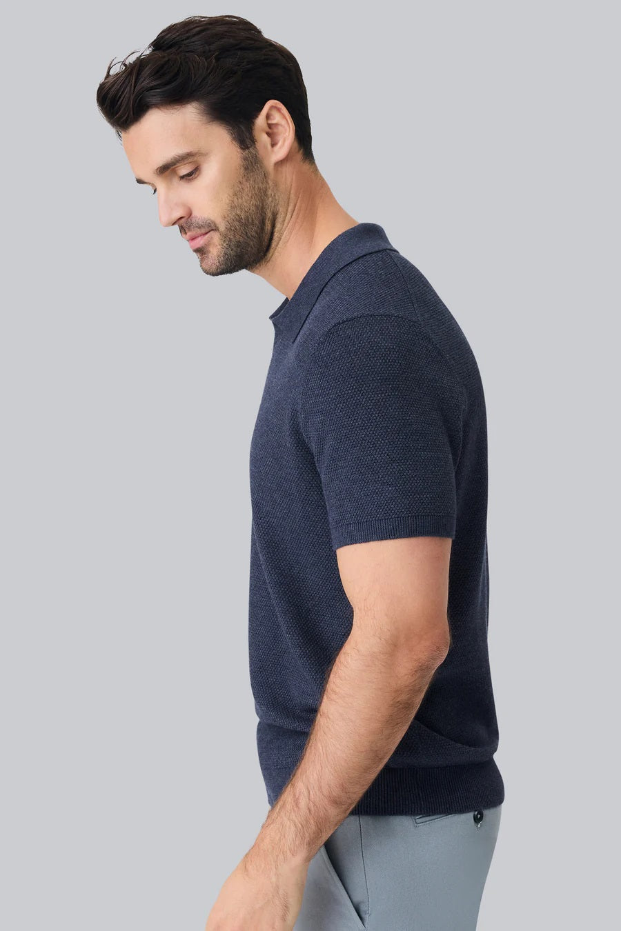 A man with short dark hair and a trimmed beard models the MACK WELDON Tech Linen Johnny Collar Polo Sweater in Navy Heather with light gray pants, standing against a plain white background.