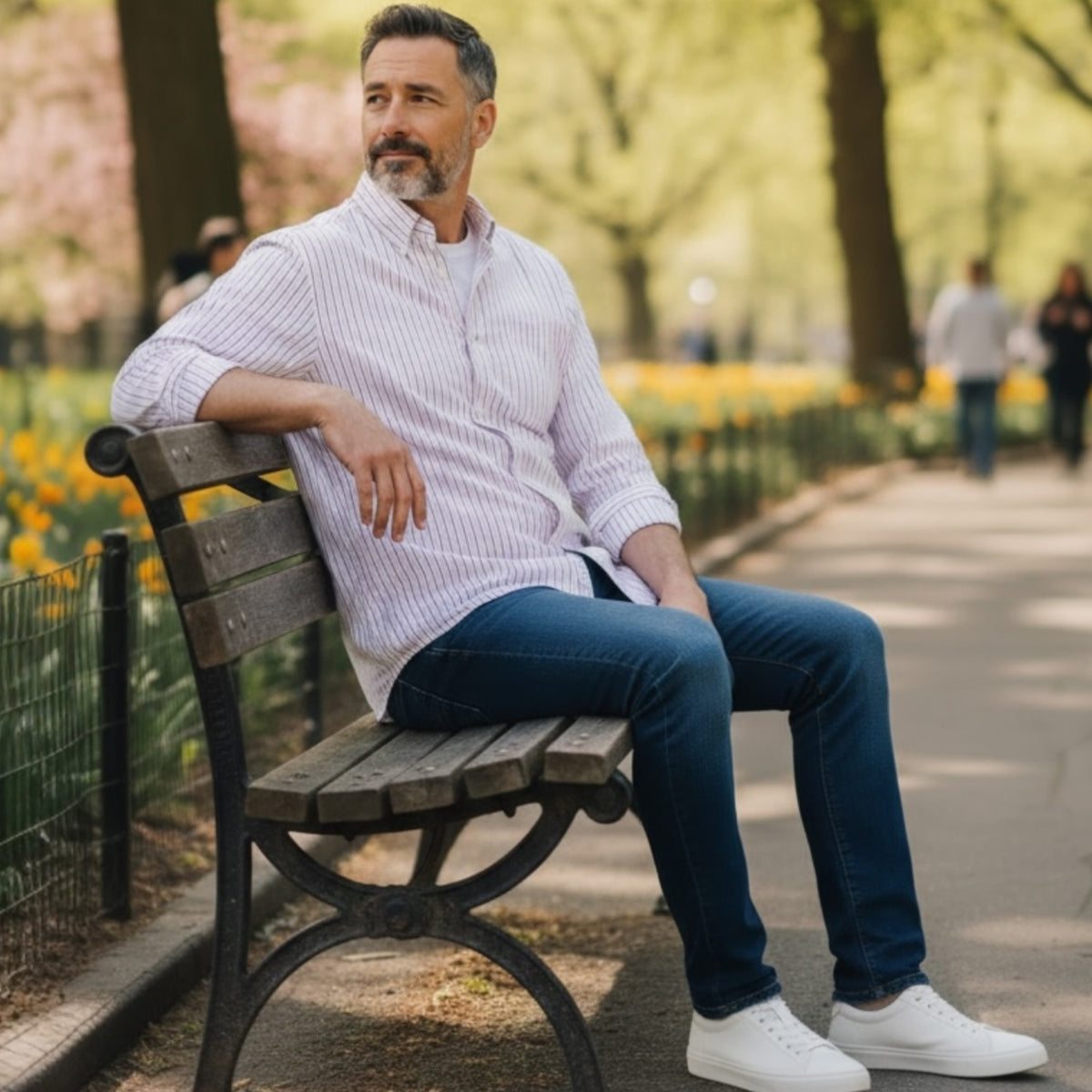 A man with short gray hair and a beard sits on a park bench wearing the FRANK & EILEEN Finbar men's button-up shirt in pink and blue multi stripe, blue jeans, and white sneakers. Behind him, people walk along a flower-lined path among trees.