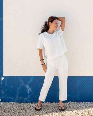 A person wears the MERSEA - FRENCHIE CIGARETTE PANT by MERSEA, paired with a white top and gray sneakers, standing on sandy beach ground with ocean waves and blue sky behind them, hands in their pockets.