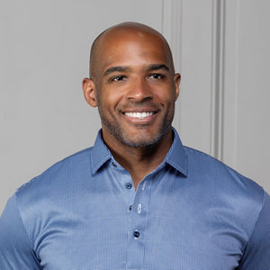 A man in sunglasses and a COLLARS AND CO - Semi Spread Collar Polo in Navy Houndstooth stands outdoors by a stone building and green plants, gazing to the side with one hand in his pocket.