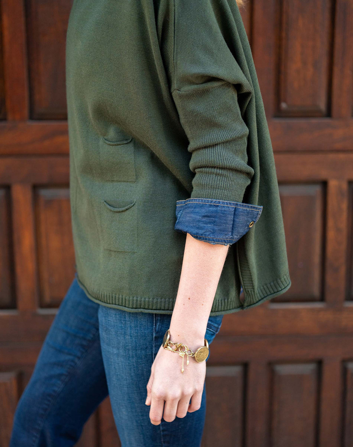 A woman with light brown hair stands before a wooden door, wearing the MERSEA - CATALINA CREWNECK SWEATER IN DEEP EMERALD over a denim shirt and blue jeans, gazing to the side with hands in her pockets and a relaxed expression.