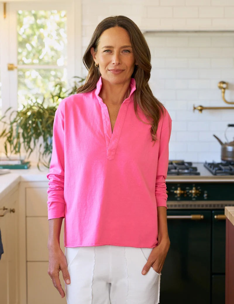 A woman with long brown hair stands in a sunlit kitchen, wearing the FRANK &amp; EILEEN Patrick Popover Henley in Lotus and white pants. Light pours through a window adorned with plants, capturing California Heritage Collection vibes.