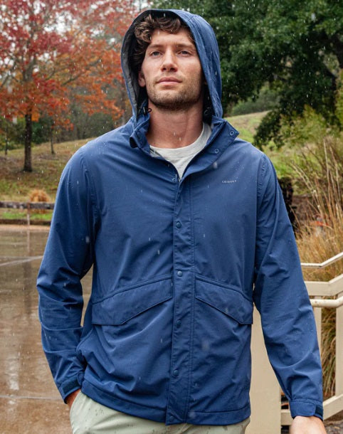 A smiling man with gray hair and a beard wears the CRIQUET Watershed Rain Jacket in Navy over a white shirt and beige pants, standing against a plain white background.