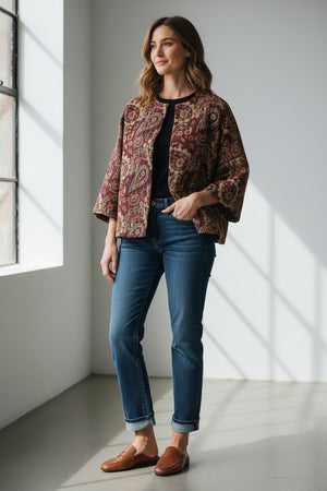 A woman stands against a plain background wearing the MIRTO 1956 Burgundy Paisley Jacquard Jacket—an oversized, cotton-blend piece in rich red and purple tones from MIRTO 1956—styled over a long burgundy dress and black shoes. The relaxed fit of the jacket complements her straight dark hair and neutral expression.