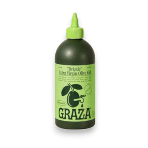 A green squeeze bottle labeled "DRIZZLE OLIVE OIL" by GRAZA sits on a gray surface against a light gray textured background. The bottle features green and black graphics, including an olive branch, making it perfect for adding flavor as a finishing oil.