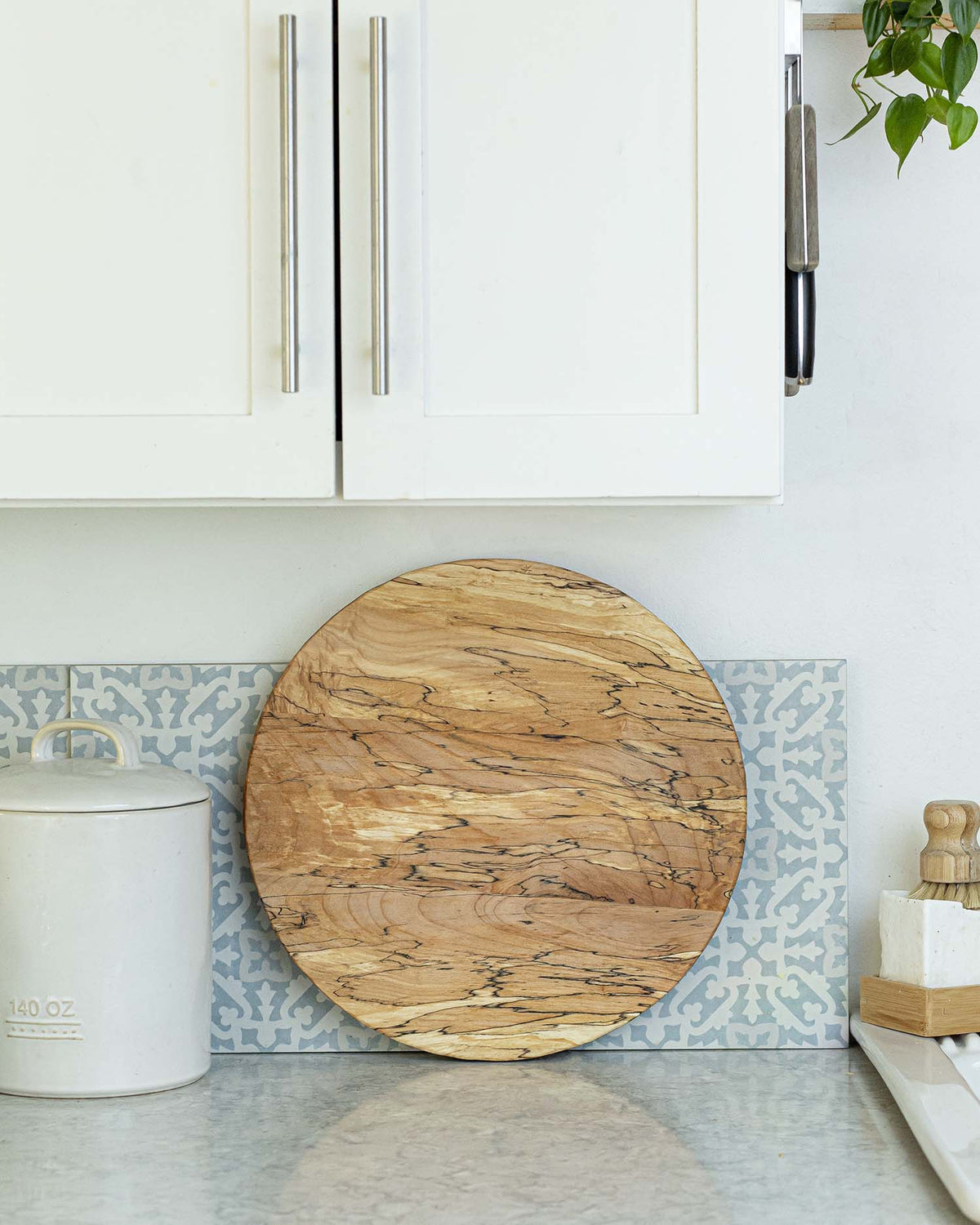 A small white vase filled with yellow and white daisy flowers sits on a PETERMANS BOARDS &amp; BOWLS INC SPENCER PETERMAN - SPALTED LAZY SUSAN 20&quot; along with a wedge of soft cheese topped with herbs and a few round crackers. The spinning tray rests on a kitchen counter, showcasing a patterned tile backsplash in the background.