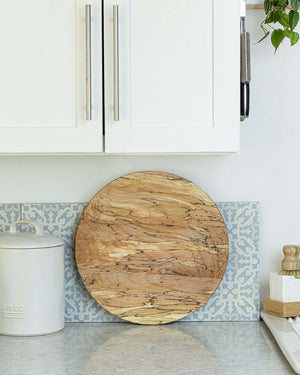 A round wooden cutting board with natural grain rests upright on a kitchen counter, perfect paired with the PETERMANS BOARDS & BOWLS INC SPENCER PETERMAN - SPALTED LAZY SUSAN 20" for extra artisan charm.