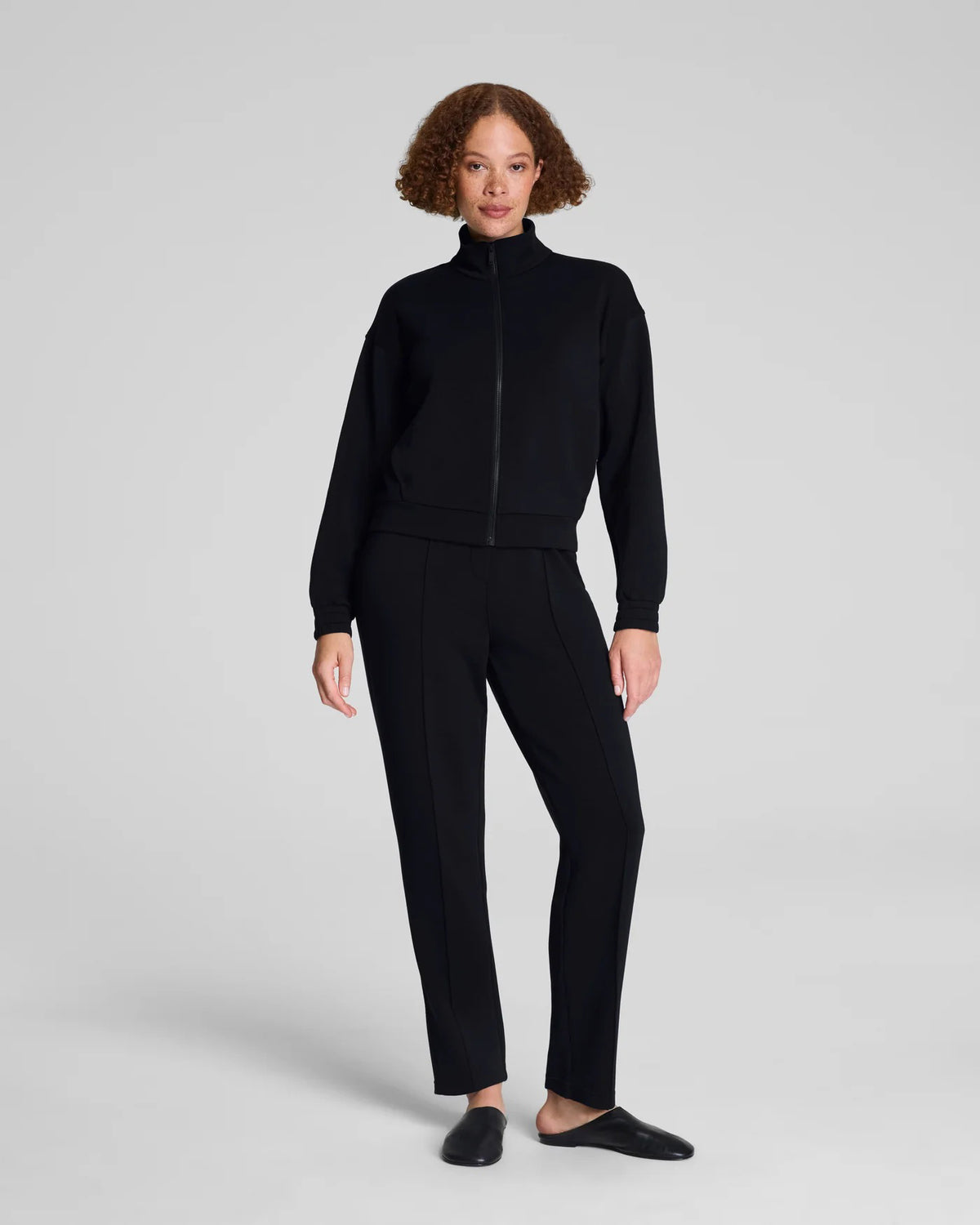 A person models the SPANX - AIRessentials Brushed Slim Straight Pant with an elastic waistband and buttons, paired with black slip-on shoes against a plain light background.