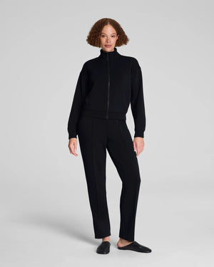 A person models the SPANX - AIRessentials Brushed Slim Straight Pant with an elastic waistband and buttons, paired with black slip-on shoes against a plain light background.
