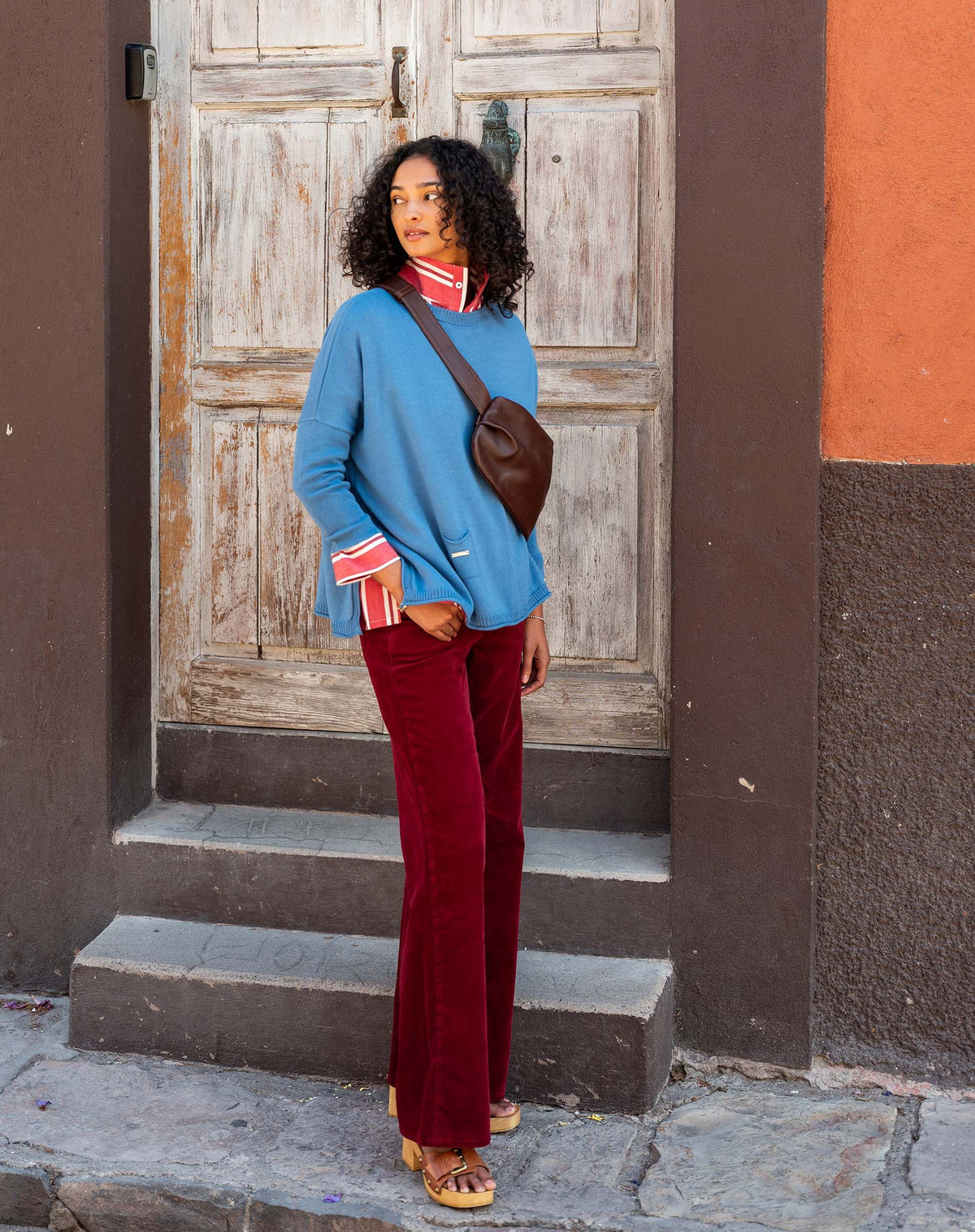 A person with curly hair walks along a two-toned wall, wearing the MERSEA - CATALINA CREWNECK SWEATER IN BLUE DUSK by MERSEA over a red collared shirt and red pants, looking down with a calm expression.