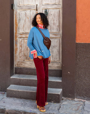 A person with curly hair walks along a two-toned wall, wearing the MERSEA - CATALINA CREWNECK SWEATER IN BLUE DUSK by MERSEA over a red collared shirt and red pants, looking down with a calm expression.