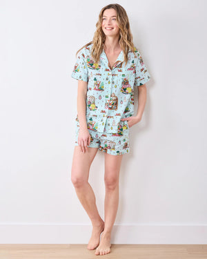 A woman with long blonde hair stands by a shelf of books in a cozy, well-lit room, wearing the PRINTFRESH - READING NOOK PAJAMA SHORT SET by PRINTFRESH—a matching light blue organic cotton short-sleeve top and pajama shorts adorned with colorful printed illustrations.