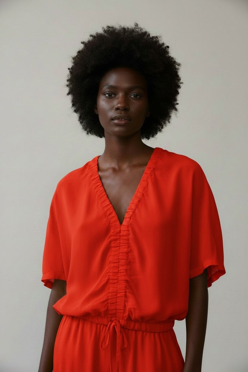 A woman with a natural afro wears the NUDE V Neck Tie Waist Blouse by NUDE, featuring a deep V-neck and short sleeves, styled in a relaxed silhouette against a plain light background.