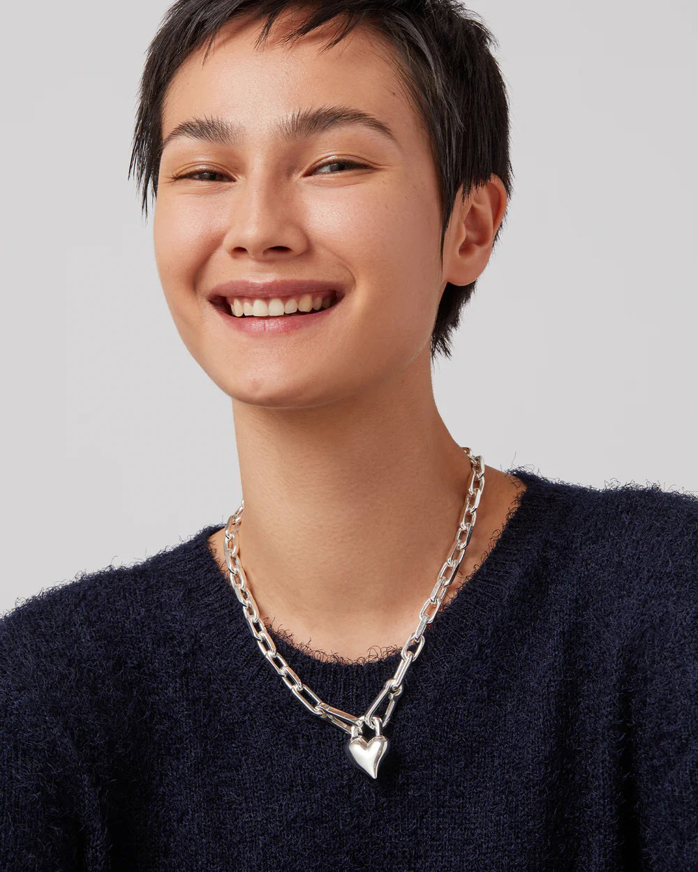 The JENNY BIRD HEART PENDANT NECKLACE showcases faceted gold chain links with a smooth, shiny heart charm at the center, elegantly set against a white background.