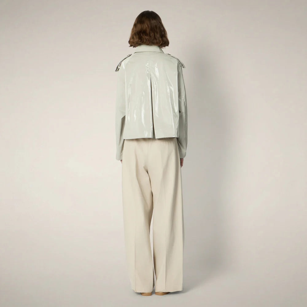 A person with chin-length brown hair wears the SAVE THE DUCK - GAIA JACKET by SAVE THE DUCK, a shiny light-gray, cropped trench-style women's jacket over a white top, paired with high-waisted beige trousers and beige shoes against a plain background.