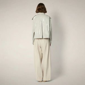 A person with chin-length brown hair wears the SAVE THE DUCK - GAIA JACKET by SAVE THE DUCK, a shiny light-gray, cropped trench-style women's jacket over a white top, paired with high-waisted beige trousers and beige shoes against a plain background.