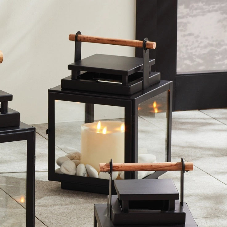 The NAPA HOME AND GARDEN - ADWIN OUTDOOR LANTERN is a black metal lantern with clear glass panels and a unique design, featuring a teak handle. It holds an unlit white candle inside, making it ideal for outdoor use with its contemporary and minimalist style.