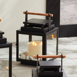 The NAPA HOME AND GARDEN - ADWIN OUTDOOR LANTERN is a black metal lantern with clear glass panels and a unique design, featuring a teak handle. It holds an unlit white candle inside, making it ideal for outdoor use with its contemporary and minimalist style.