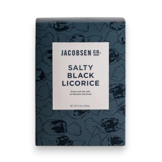 A dark blue JACOBSEN SALT - SALTY BLACK LICORICE BOX by JACOBSEN SALT CO. rests on a light surface against a plain gray background. Its white label with black text and abstract patterns hints at the sea salt and anise flavors inside.