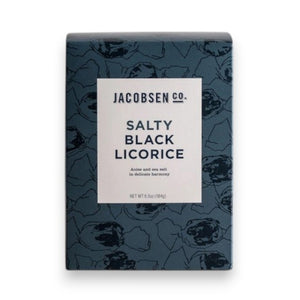 A dark blue JACOBSEN SALT - SALTY BLACK LICORICE BOX by JACOBSEN SALT CO. rests on a light surface against a plain gray background. Its white label with black text and abstract patterns hints at the sea salt and anise flavors inside.