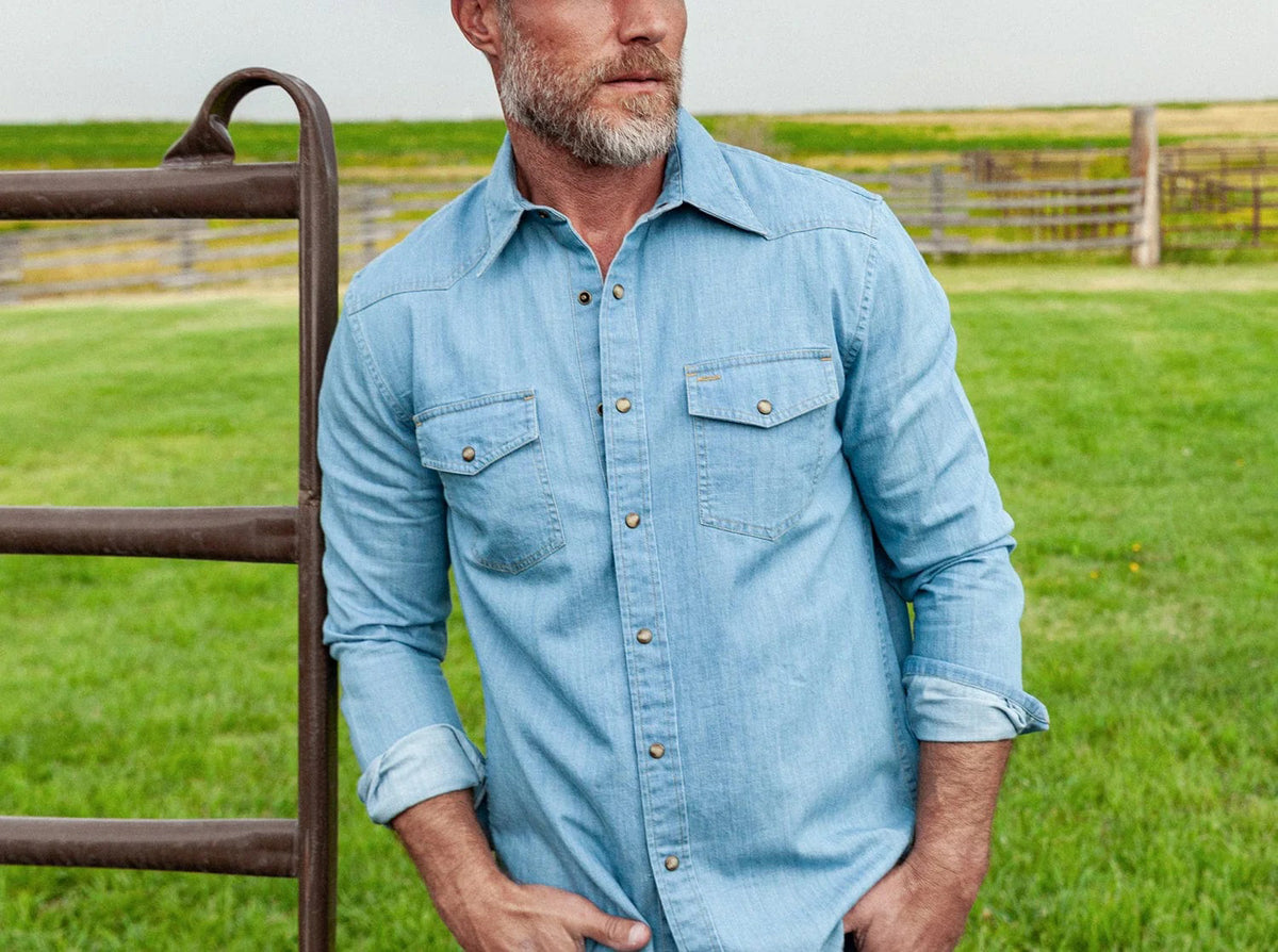 A man with a gray beard wears the CRIQUET WESTERN DENIM SHIRT WITH A PEARL SNAP IN LIGHT WASH by CRIQUET as he stands outdoors near a metal fence, with green grass and an open field in the background.