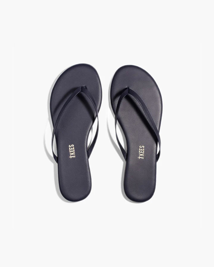 The TKEES - LILY LINER SLIPPER TWILIGHT by TKEES INC are simple dark flip-flops with thin straps and cushioned insoles, featuring the brand name printed on each insole and displayed on a white background.