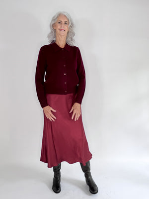 An older woman with long gray hair stands smiling in a dark red ROSE & CROWN cardigan, the ROSE AND CROWN - SATIN MIDI SKIRT, and black knee-high boots against a plain white background.